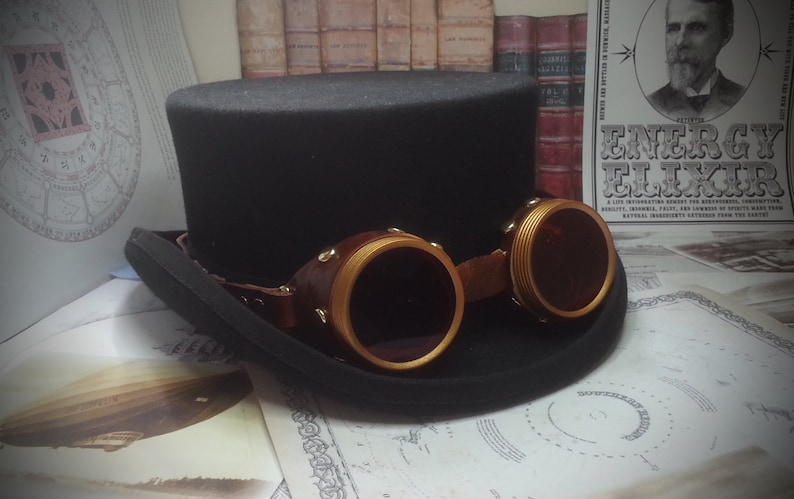 May include: A black top hat with a wide brim and a pair of gold-rimmed goggles with brown leather straps. The goggles have round lenses and are attached to the hat with a strap.