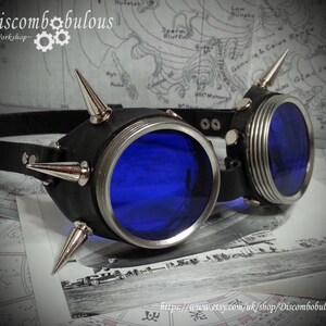 May include: A pair of steampunk goggles with blue lenses and a black leather frame. The frame is decorated with silver spikes. The goggles are lying on a map.