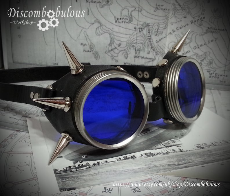May include: A pair of steampunk goggles with blue lenses and a black leather frame. The frame is decorated with silver spikes. The goggles are lying on a map.