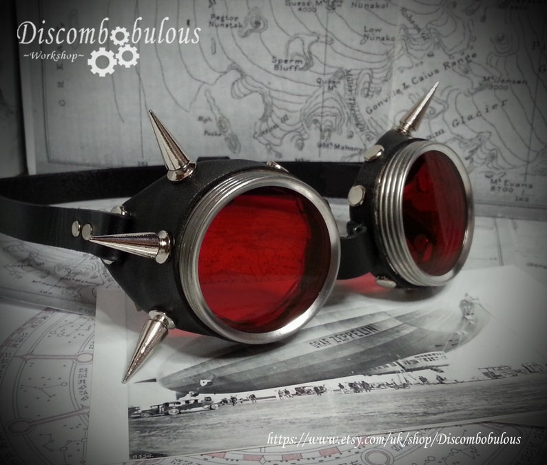 May include: A pair of steampunk goggles with red lenses and silver accents. The goggles are adorned with spikes and have a black leather strap.