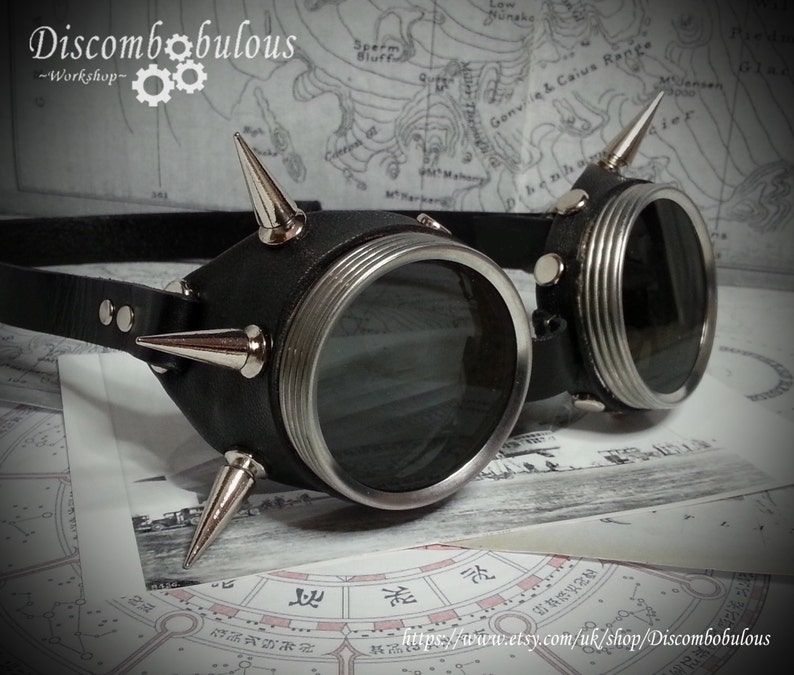May include: Black leather steampunk goggles with silver accents and spikes. The goggles have round lenses and a black leather strap.