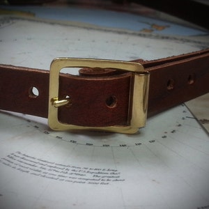 May include: A brown leather belt with a gold metal buckle. The belt has two rows of stitching and a single hole on each side of the buckle.