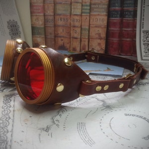 May include: A pair of steampunk goggles with red lenses and a brown leather strap. The goggles have gold accents and rivets.