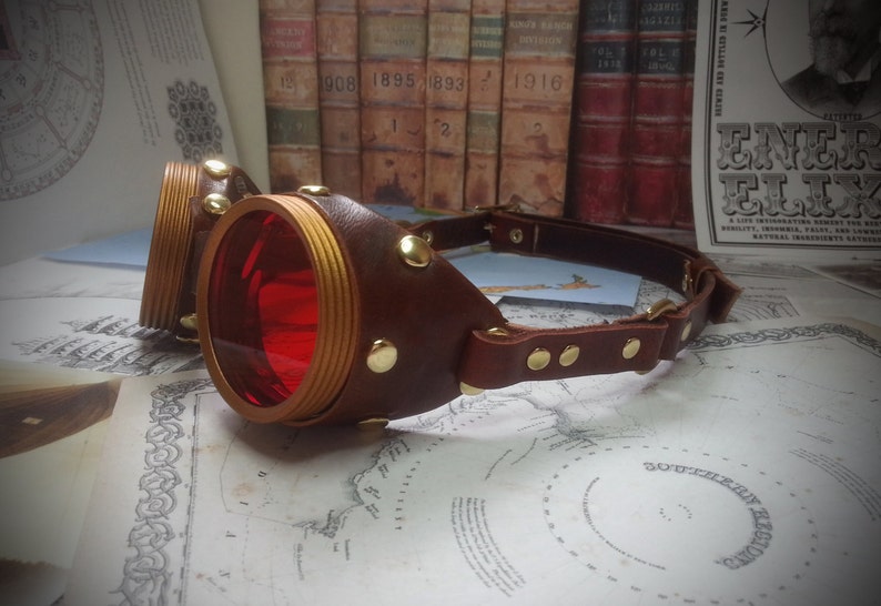 May include: A pair of steampunk goggles with red lenses and a brown leather strap. The goggles have gold accents and rivets.