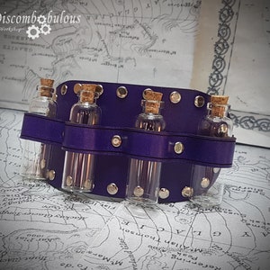 May include: A purple leather wrist cuff with six small glass bottles with cork tops. The bottles are arranged in a row and are held in place by the leather cuff. The cuff is decorated with metal studs.