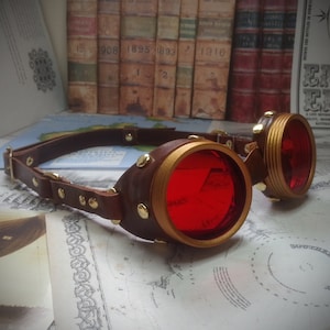 May include: A pair of steampunk goggles with a brown leather strap and red glass lenses. The lenses are round and have a gold metal frame.