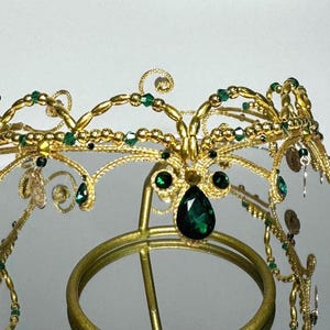 Może przedstawiać: Złota korona z misterną drucianą pracą, ozdobiona zielonymi kamieniami szlachetnymi i małymi złotymi akcentami. Korona ma duży, łezkowaty szmaragdowy klejnot pośrodku i jest prezentowana na złotym stojaku.
