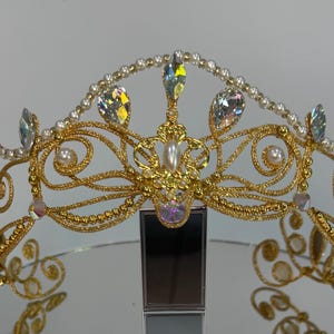 Puede incluir: Una tiara dorada con un diseño ornamentado. La corona presenta piedras preciosas claras e iridiscentes, perlas y un elemento decorativo central. La tiara se exhibe sobre un soporte contra un fondo neutro.
