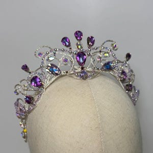 May include: A silver tiara with a delicate filigree design, adorned with large, teardrop-shaped purple gemstones and smaller iridescent crystals. The tiara is set on a neutral-toned mannequin head, showcasing its intricate details and sparkling embellishments.