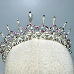 May include: A silver-toned crown with a delicate design. The crown features white faux pearls, pink and clear rhinestones, and teardrop-shaped accents. The crown is displayed on a mannequin head.