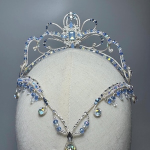 Puede incluir: Una tiara plateada con detalles de cristal azul y transparente. La corona presenta un cristal central de corte cuadrado y un diseño delicado y fluido. Un colgante a juego cuelga debajo, lo que se suma a la estética elegante y de cuento de hadas.