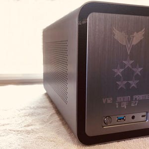 Op de afbeelding: Een donkergrijze computerkast met een geborsteld metalen voorpaneel. Het paneel heeft een geëtst ontwerp van een vogel en sterren, met de tekst "V12 JOHN PRIME 1 OF 27". De kast heeft een USB-poort en een aan/uit-knop.