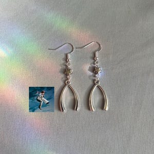May include: A pair of silver-colored earrings. Each earring features a fish hook, a small bead, a star-shaped charm, and a wishbone-shaped pendant. The background is a light gray fabric with a rainbow-colored light effect.