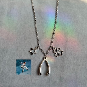 May include: Silver-toned necklace with a wishbone, star, and four-leaf clover charms. The chain hangs on a light gray surface with a rainbow light effect. A small image of a person skydiving is in the lower left corner.