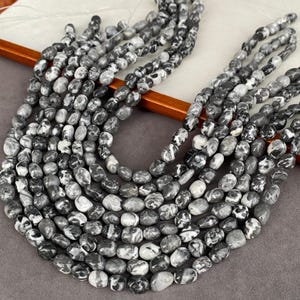 Puede incluir: Múltiples hilos de cuentas de forma ovalada con un patrón jaspeado en tonos negro, blanco y gris. Cada cuenta tiene una superficie lisa y pulida con texturas arremolinadas únicas, ensartadas en hilos de filamento finos, claros y flexibles.