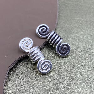 Könnte beinhalten: Zwei Haarspiralen aus silberfarbenem Metall, die jeweils ein zentrales Spiraldesign mit einem gewickelten Drahtkörper aufweisen. Die Haarschmuckstücke haben eine polierte Oberfläche und sind als kleine Zylinder mit dekorativen Wirbeln geformt.