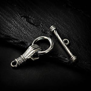 Puede incluir: Un conjunto de cierre de palanca plateado que consta de un anillo circular con un detalle de mano esculpida y una barra recta a juego con un pequeño bucle central, ambos con un acabado oxidado y texturas metálicas detalladas.