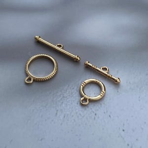Può includere: Due chiusure a T dorate per la creazione di gioielli. Ogni set è composto da un anello circolare con un piccolo occhiello alla base e una barra a T abbinata. Un anello presenta una texture a corda intrecciata, l'altro segmenti lisci.