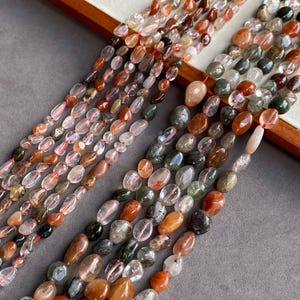 Puede incluir: Múltiples hilos de cuentas pulidas de forma ovalada irregular en tonos transparente, blanco translúcido, rojo intenso, naranja quemado y verde bosque oscuro. Las cuentas están ensartadas en un hilo rojo fino, mostrando inclusiones.