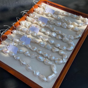 Könnte beinhalten: Fünf Stränge unregelmäßiger, cremefarbener Barockperlen werden auf einem Holztablett präsentiert. Jeder Strang ist mit einem Buchstaben von A bis E gekennzeichnet. Die Perlen haben einen glänzenden Glanz und variieren in Größe und Form, was ihre natürliche Schönheit zeigt.