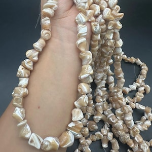 Könnte beinhalten: Eine Ansammlung unregelmäßiger, getrommelter Muschelperlen in Beige-, Creme- und Brauntönen mit perlmuttartigem Glanz. Die Perlen sind auf dünnem weißem Faden aufgereiht und weisen glatte, polierte Oberflächen sowie vielfältige organische Formen auf.