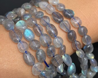 May include: Close-up of multiple strands of labradorite gemstone bracelets. The oval-shaped beads are primarily gray with iridescent blue and green flashes. The beads are strung on a thin, dark blue cord, creating a layered effect. The bracelets are displayed on a light skin tone.