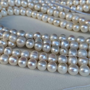 Può includere: Primo piano di più fili di perle bianco crema. Le perle sono per lo più rotonde con una lucentezza morbida. L'immagine si concentra sulla consistenza e sul colore delle perle, mostrando le loro forme individuali e il modo in cui riflettono la luce.