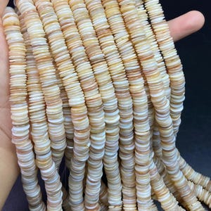 Può includere: Primo piano di più fili di piccole perline rotonde e piatte nei toni crema, beige e marrone chiaro. Le perline sono infilate insieme, creando un effetto strutturato. Le perline sono tenute in mano, suggerendo che sono per la creazione di gioielli.