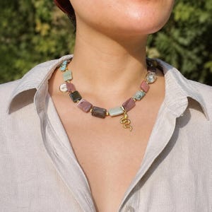 Op de afbeelding: Een ketting met rechthoekige kralen in tinten mauve, saliegroen en antracietgrijs, geaccentueerd met kleine goudkleurige bolletjes. Een goudkleurige slangenbedel hangt in het midden en een heldere, ronde gefacetteerde kristalkraal zit rechts.