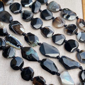 Può includere: Primo piano di diversi fili di perline di pietre preziose. Le perline sono principalmente nere con variazioni di grigio, bianco e marrone. Ogni perla ha una forma irregolare e sfaccettata, infilata con un cordoncino nero. Le pietre preziose hanno una finitura lucida e lucente.