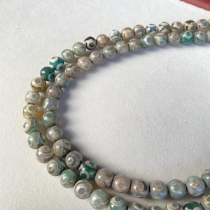 May include: Strands of round, patterned beads in shades of green, gray, and brown. The beads have a unique eye-like design, creating a natural and earthy aesthetic. The beads are strung together, suggesting a necklace or bracelet.