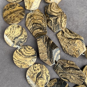May include: A collection of polished stone beads featuring a distinct marbled pattern in shades of tan, brown, and black. The beads are shaped into a mix of circular and irregular pentagonal forms, each displaying unique swirling organic textures.