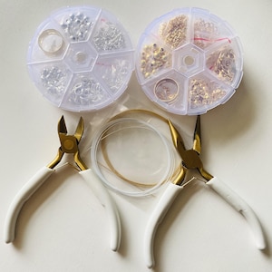 May include: Jewelry making supplies arranged on a white surface. The image features two clear, multi-compartment containers filled with silver and gold-toned jewelry findings. White-handled pliers and wire are also included, perfect for crafting.