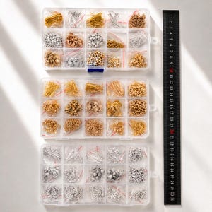 Puede incluir: Tres cajas organizadoras de plástico transparente llenas de diversos suministros para la fabricación de joyas. Las cajas contienen pequeñas bolsas de cuentas, cadenas y hallazgos de color oro, plata y cobre. Una regla negra con marcas blancas está a la derecha.