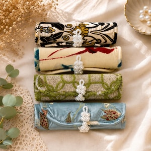 May include: Four cylindrical fabric jewelry rolls stacked vertically. Each roll features a different pattern and color, including floral and geometric designs, and is secured with a white faux pearl beaded toggle closure at the center.