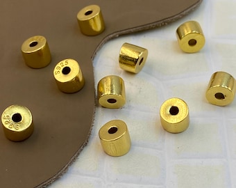 May include: A collection of small, cylindrical gold-colored beads with a central hole. Several of the beads feature the text S925 stamped on the flat top surface. The beads have a smooth, metallic finish and are scattered across the frame.