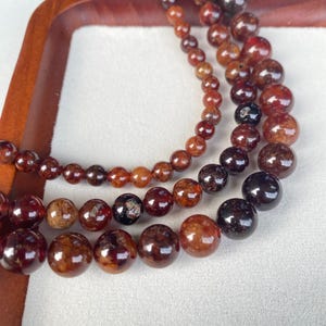 May include: Close-up of three strands of gemstone bead bracelets. The beads are various sizes and shades of brown and red. The bracelets are displayed in a wooden tray.