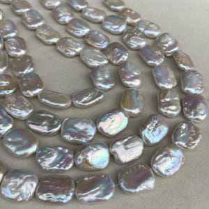 Può includere: Primo piano di fili di perle bianche d'acqua dolce. Le perle sono di forma irregolare e hanno un aspetto piatto, simile a una moneta. Mostrano colori iridescenti, tra cui rosa, verde e blu. Le perle sono infilate, per una collana o un braccialetto.