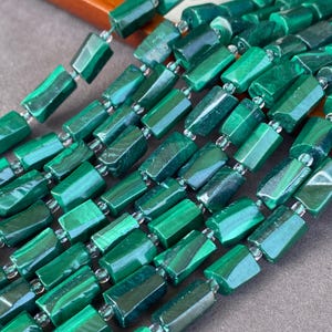 Könnte beinhalten: Eine Ansammlung rechteckiger Malachitperlen, die in mehreren parallelen Strängen angeordnet sind. Jede Perle zeigt eine tiefgrüne Farbe mit natürlichen, wirbelnden Streifenmustern und einer polierten Oberfläche, getrennt durch kleine, klare Zwischenperlen.