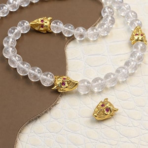 May include: A beaded necklace featuring translucent, clear spherical beads interspersed with gold-toned metal devil mask charms. Each mask charm is detailed with small, vibrant pink gemstone eyes. An additional loose devil mask charm sits nearby.
