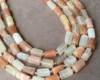 May include: A multi-strand necklace featuring cylindrical beads in varying shades of peach, cream, and translucent white. The polished stone beads are strung together in a layered arrangement, showcasing a smooth, semi-opaque texture throughout.