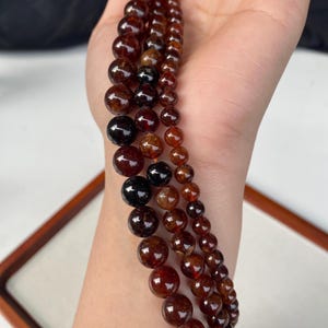 May include: Three strands of polished, dark red garnet beads of varying sizes. The beads range from small to large, with a deep, rich color. The beads are strung together, creating a layered effect. The background is a neutral color.