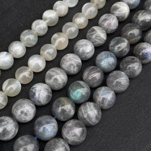 Puede incluir: Primer plano de varios hilos de cuentas de piedras preciosas. Las cuentas son redondas y varían en color, incluyendo blanco, gris y tonos iridiscentes. Las cuentas están ensartadas, mostrando sus superficies pulidas y variaciones naturales. Ideal para la fabricación de joyas.