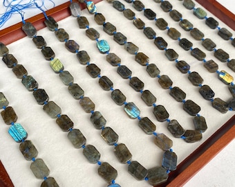 May include: Strands of labradorite gemstone beads. The beads are a mix of gray, brown, and blue hues, with flashes of iridescent color. Each bead is a rectangular shape, strung on blue thread. The beads are displayed in a wooden tray.