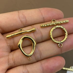 May include: Two gold-toned toggle clasps for jewellery making. One set features a smooth, irregular bar and an organic, rounded loop. The second set includes a textured, hammered bar and a matching circular ring with a hammered surface finish.