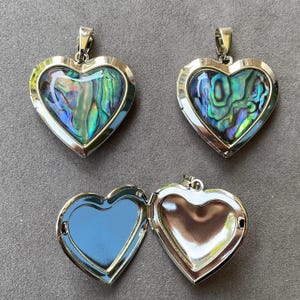 Pode incluir: Três relicários em forma de coração feitos de metal prateado polido. Dois relicários estão fechados, exibindo incrustações de concha de abalone iridescente com padrões de redemoinhos verdes, azuis e roxos. Um relicário está aberto, revelando um interior liso.