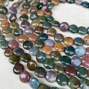 Op de afbeelding: Close-up van strengen gepolijste edelsteen kralen in verschillende kleuren, waaronder groen, bruin, paars en oranje. De platte, ronde kralen hebben een glanzende afwerking. Geschikt voor het maken van sieraden.