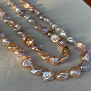 Könnte beinhalten: Drei Stränge aus glänzenden, unregelmäßig geformten Süßwasserperlen in den Farbtönen Pfirsich, Creme und schillerndem Weiß. Die Perlen sind zu einer Halskette aufgefädelt. Die Perlen haben eine glatte, polierte Oberfläche.