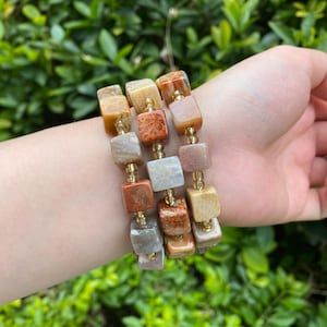 Peut inclure: Trois bracelets de perles avec des pierres carrées multicolores dans les tons beige, orange et gris. Les perles sont enfilées sur un fil doré. Les bracelets sont portés sur un poignet, avec un fond de feuilles vertes.