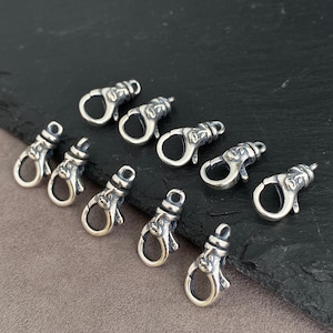 Puede incluir: Un conjunto de ocho cierres de mosquetón de metal plateado, cada uno con un diseño de rostro detallado en relieve en el cuerpo principal. Estos fornituras para joyería tienen un acabado pulido con áreas oscurecidas para resaltar los rasgos.